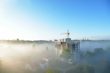 Construction cranes on a background of blue foggy sky. The work of construction equipment. Cranes work in the fog