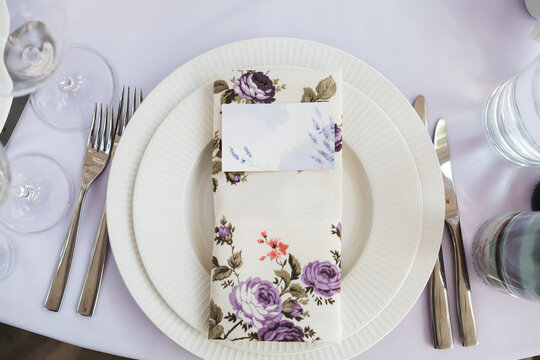 Top view of plates anf cutlery with serviette with violet flowers print anf tablet with guest name on the white table, restaurant table serving, preparing for the wedding celebration