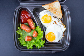 Fried eggs with sausages and croutons on a dark background. Scrambled eggs in a disposable plate close-up.