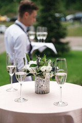 Front view of three champagne glasses full with sparkling drink on table, floral composition made of eustomas and blurred waiter on background. Luxury wedding catering. Concept of modern celebrating.