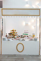 Full view of white candy bar table full with baked sweets and fruits. Decorated elegant candy cart. Luxury wedding catering, party table serving. Concept of modern decorated party celebration.