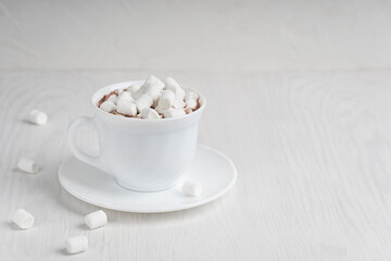 White cup full of hot cacao drink with marshmallow looking as snow usually cooked at cold winter and autumn days served on round plate on wooden background. Image with copy space, horizontal