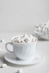 White cup full of warm sweet homemade cacao drink usually prepared at cold winter and autumn sesons served on plate with bowl of marshmallows on wooden background. Vertical orientation image