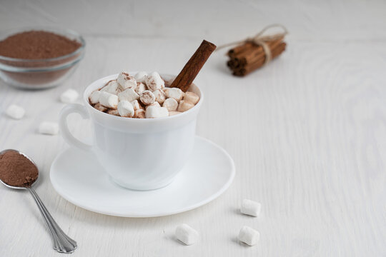 Cup full of hot homemade cacao drink usually prepared at cold winter days served on plate with marshmallows, cinnamon and spoon on white wooden background at kitchen. Image with copy space, horizontal