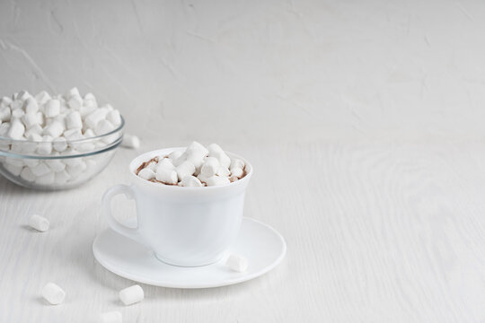 Hot sweet homemade cacao drink usually prepared at cold autumn and winter days in white cup standing on plate with marshmallow in glass bowl on wooden background, Image with copy space, horizontal