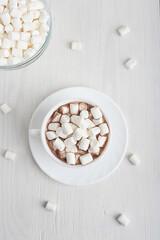 View from above on cup full of sweet homemade cocoa drink usually prepared at cold winter vacations served on plate with bowl of marshmallow on white wooden background. Vertical orientation image