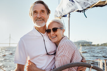 Cheerful senior couple standing at steering wheel on a yacht and hugging each other. Happy people...