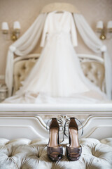 Close up of fashionable shoes on high heel and blurred wedding dress on background. Beautiful bridal clothing elements. Luxury footwear for important formal occasion. Concept of old fashioned style.