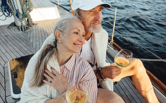 Senior Couple Wrapped With Plaid And Sitting On A Yacht Deck With Bocals Of Wine. Two Loving Mature People Sitting Together On A Sailboat And Looking Away.