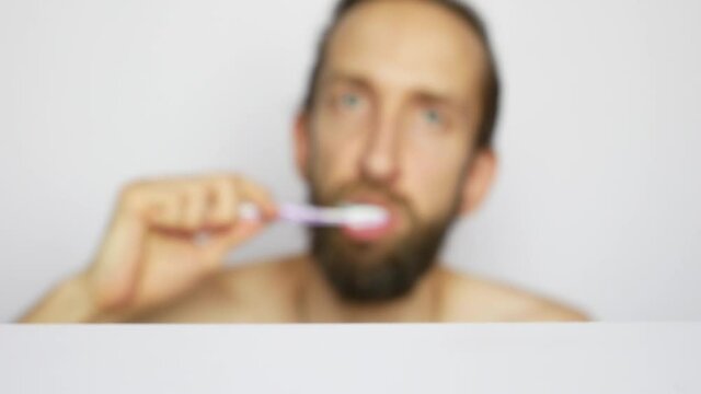 A young bearded man takes a toothbrush from the shelf and brushes his teeth