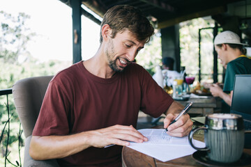 Young male freelancer making notes on information during remote job on cafe terrace, smiling handsome hipster guy writing and filling in application for employment using pen and documentation