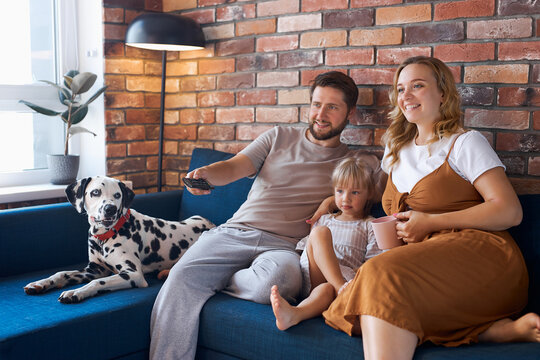 Attractive Happy Family With Dog Watch Tv In Living Room, Young Man, Woman, Daughter And Pet Dog Sit On Sofa, Weekends