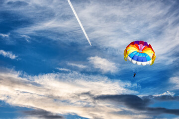 A jet flying high in the blue sky with a white trace behind and a parachute gliding under it.