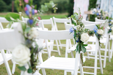 Close up stylish white chiavari chair for guest on wedding ceremony decorated with white rose, blue flower, greenery. Element of furniture for elegant open air event. Concept of tenderness in design.