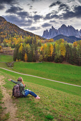 Fototapeta na wymiar Girl with backpack is in famous best alpine place of the world .Santa Maddalena village with Dolomites mountains in background, Val di Funes valley, Trentino Alto Adige region, Italy, Europe