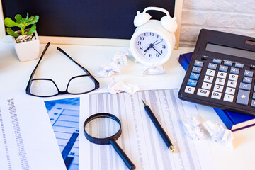 An office table with a magnifying glass, diaries, calculator, white alarm clock, pencils, and a whiteboard stands against a light brick wall. Workplace top view. Business concept