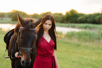 Fototapeta premium A young pretty girl in a red dress poses on a ranch with a thoroughbred stallion at sunset. Love and care for animals