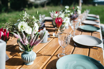 Close up of blue plates on wooden table, tender bouquets on backyard open air wedding celebration. Birthday party on sunny day outdoors. Decoration and table serving. Concept of catering tenderness.