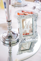 Close up of table number four on reflecting glass table and silver candlestick. Preparation for elegant gala night, prom. Details in restaurant interior. Concept of design elements and classy motives.