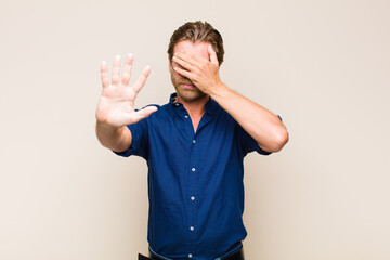 blonde adult caucasic man covering face with hand and putting other hand up front to stop camera,...