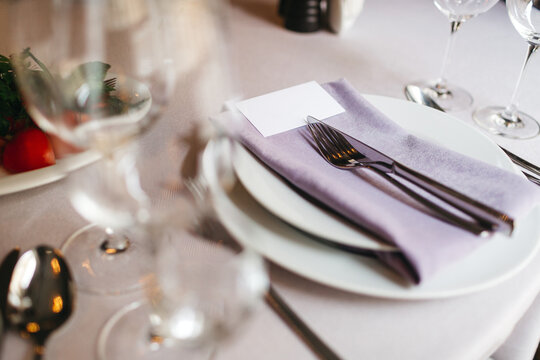 Crop of served table with plates, violet serviette, silver cutlery, blurred vine glasses on foreground. Elegant catering of beautiful restaurant, wedding hall preparing. Concept of minimalism in cafe.