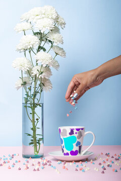 White flowers that look like cotton flakes with green stem or branch glass vase with water on a blue and pink background, posillo with colored hearts pastel chip female hand drops candy