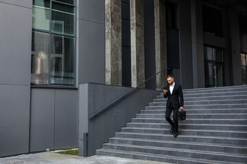 businessman walking from office building and looking at phone ou