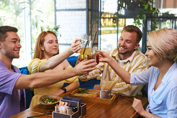 group of friends clink glasses with drinks in cafe, young men and women gathered to have fun and talk, share news