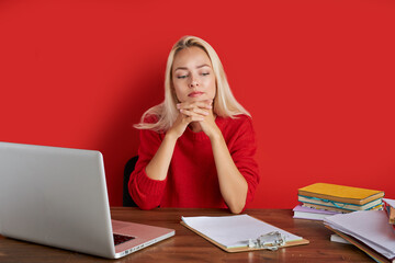 portrait of confident caucasian blonde female sitting at office desk with laptop and papers, young lady in casual wear work as manager, has too much work, think, isolated over red background