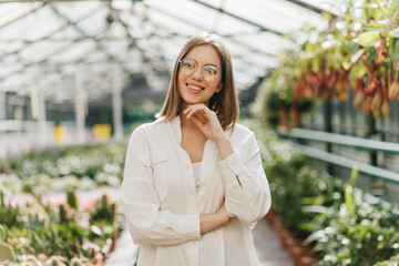 Pensive dark-haired girl standing in greenhouse. Portrait of adorable lady in white blouse posing on blur background.