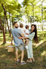 Fototapeta premium Adorable parents with little children walking at the park, caring mother hold in arms cute son, beautiful daughter sitting in father hands, happy family moments, parenting concept