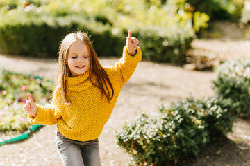 Good-humoured kid in sweater dancing on nature background. Outdoor photo of refined little girl with brown hair fooling around in park.