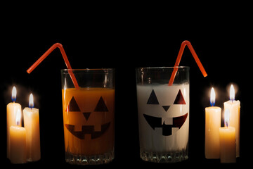 There are two glasses of white and orange drinks on a black background, lit by candles, and on the glasses are an eerie smiling face. Halloween. Idea - drinks for the holiday