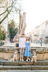 Happy Caucasian young couple in love, with two husky dogs, posing to camera in the center of ancient European city standing near old fountain on stone stairs. Couple in love with dogs in the city