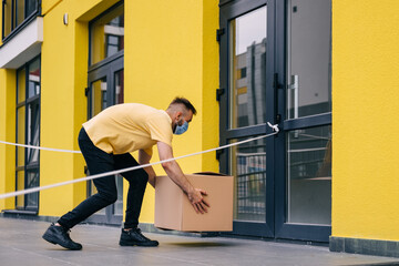 A courier wearing a medical mask safely delivers online purchases in a cardboard box to the door during the COVID-19 coronavirus epidemic. Stay home safe concept.
