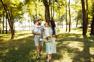 Fototapeta premium Loving parents with little kids walking at the park, adorable mother hold in arms beautiful daughter, cute son sitting in fathers hands, enjoy happy family weekend concept