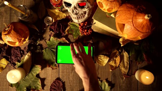 Using a phone with horizontal green screen at Halloween decorations. Top view decorations for all saints day and smartphone. Mystical tradition in harvest autumn desk decor, copy space template.