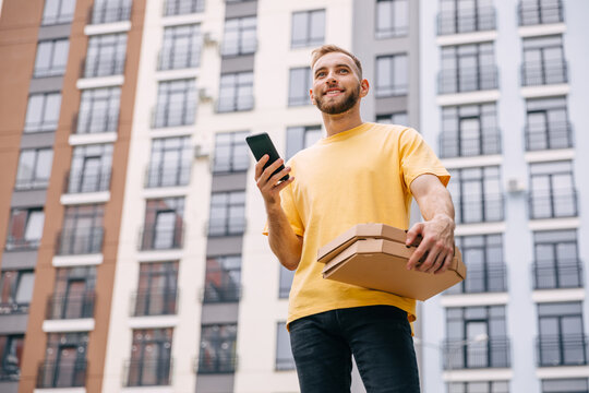 Low Angle View Of Courier Holding Pizza Boxes And Using Smartphone On Urban Street