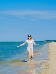 Fototapeta premium A young girl runs along the sandy sea beach along the surf. Splashes of sea water. The girl is wearing a white swimsuit and sunglasses. Freedom and carelessness. Outdoor fitness. Sunny day. Copy space