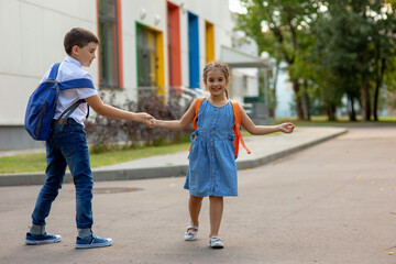a smiling little girl and a brother with backpacks hold hands, play and have fun near the school building