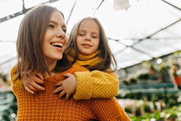 Dreamy young woman playing with daughter. Adorable smiling lady in sweater posing on orangery with kid. - obrazy, fototapety, plakaty