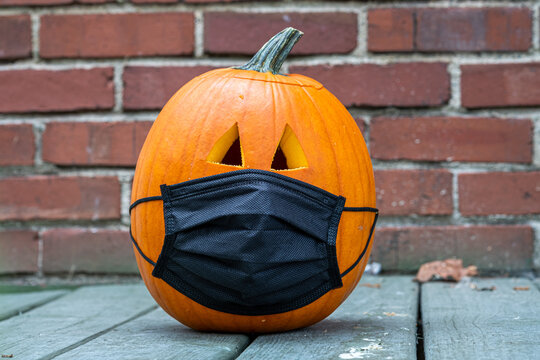 Carved pumpkin for Halloween wearing a Covid 19 face mask on a porch with a brick background