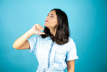 Fototapeta premium Young beautiful woman wearing a denim jumpsuit over isolated blue background with angry face, negative sign showing dislike with thumb down