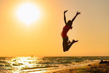 Happy girl jumping on the beach against the background of the sea and sunset. Sunset at sea. Girl jumping raising her hands up.