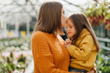 Fototapeta premium Young woman kissing daughter in forehead. Photo of lovely lady spending time with child in greenhouse.