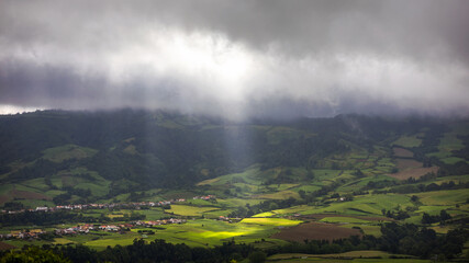 Fototapeta premium Bright sunlight shining through hole of clouds to dark scene of mountain range, Azores, Sao Miguel, Portugal. The beam of light through the dark clouds on the mountains in Azores, Portugal.
