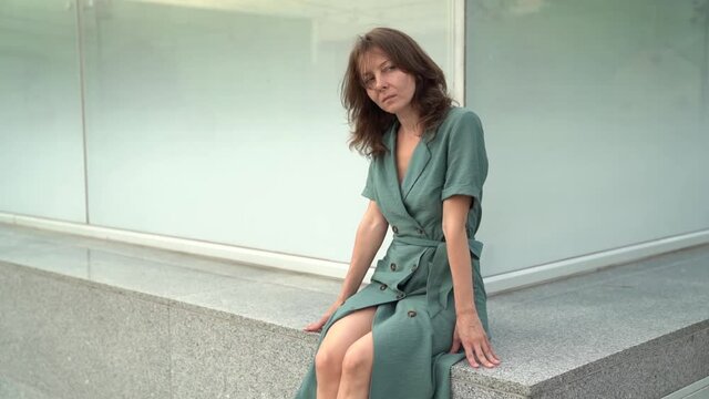 sexy middle-aged woman is posing outdoors, sitting on stone bench at modern city