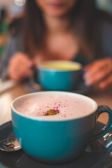 Close up on a foamy rose latte coffee with petals in a blue mug and woman in the background
