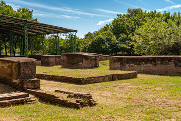 Fototapeta na wymiar Leon, Nicaragua - November 27, 2008: Park setting of low brick walls at the ruins of old Leon under blue sky with white stripes. Green lawn, green tree foliage and shade producing hangar roof.