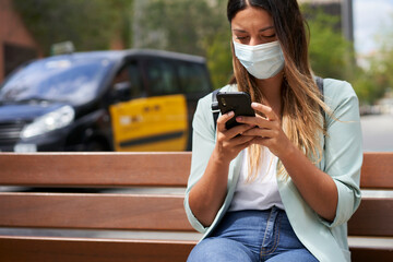 Working woman with face mask in a city, sitting on a bench, using a smartphone. Behind it is a...